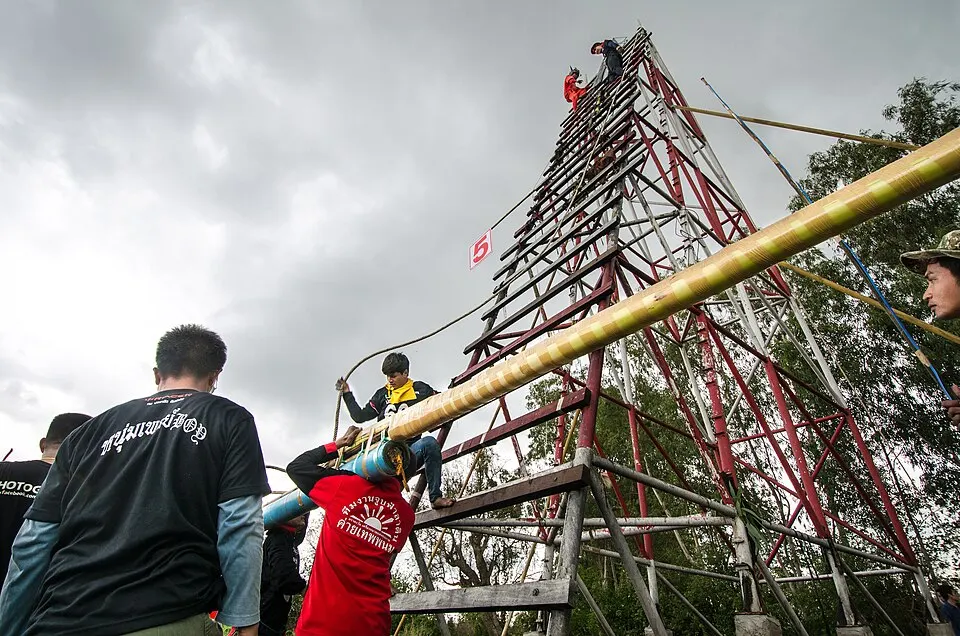 Men hauling a long bamboo rocket up a tall wooden launch tower under a grey sky